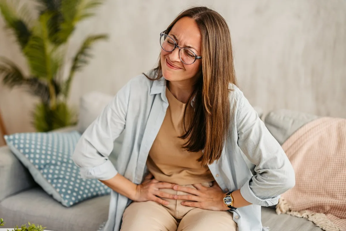 Foto colorida de mulher sentada em sofá com a mão no abdômen - Metrópoles