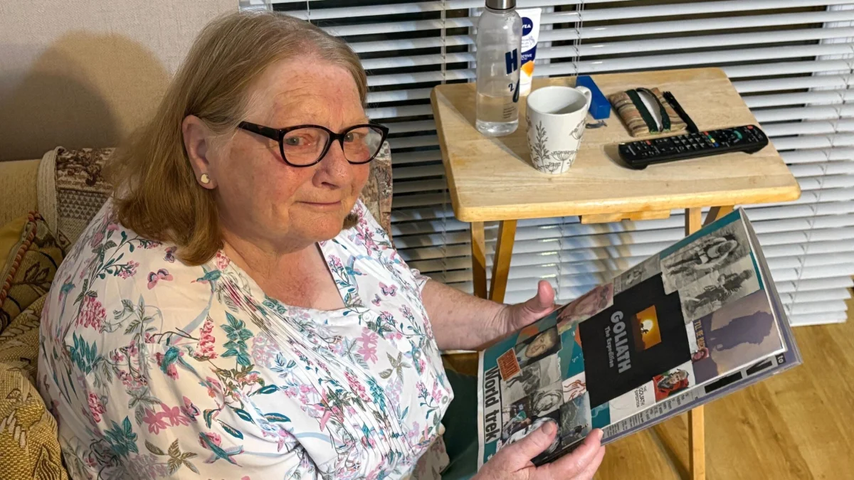 Uma mulher de óculos, com cabelos castanho-claros já grisalhos, segura um álbum de recortes com fotos do filho coladas na capa