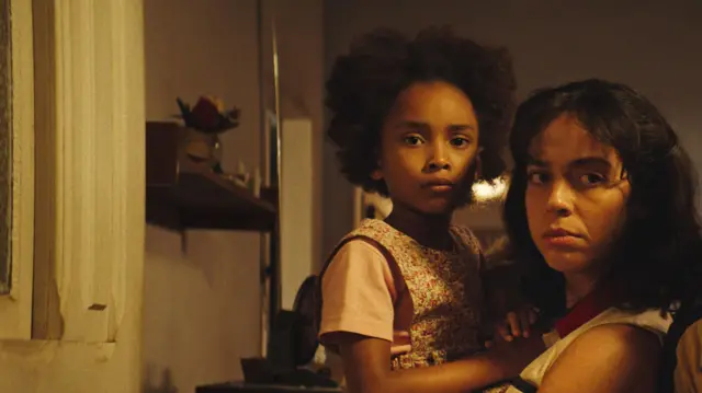 Uma mulher segura no colo uma criança em cena de Emergência Radioativa. Elas estão em uma casa com iluminação quente, próximas uma da outra, olhando em direção à câmera com expressões sérias e tensas; ao fundo, há uma parede com uma pequena prateleira e objetos.