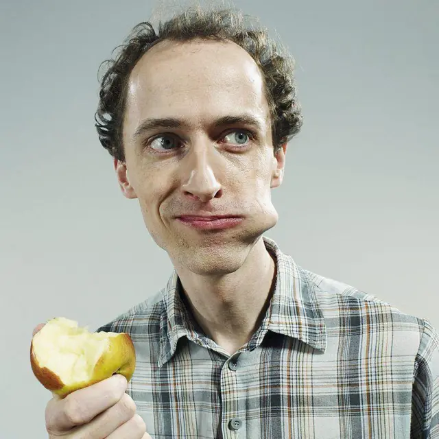 Homem comendo maçã e olhando para o lado, com um pedaço da fruta na boca